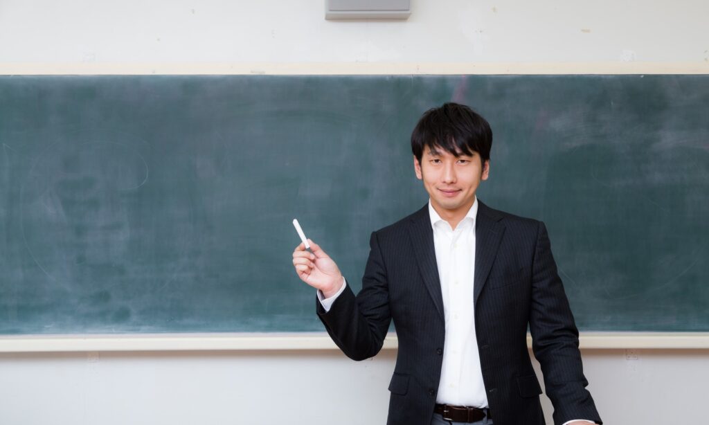 教師が白のチョークを持って、黒板の前に立っている。